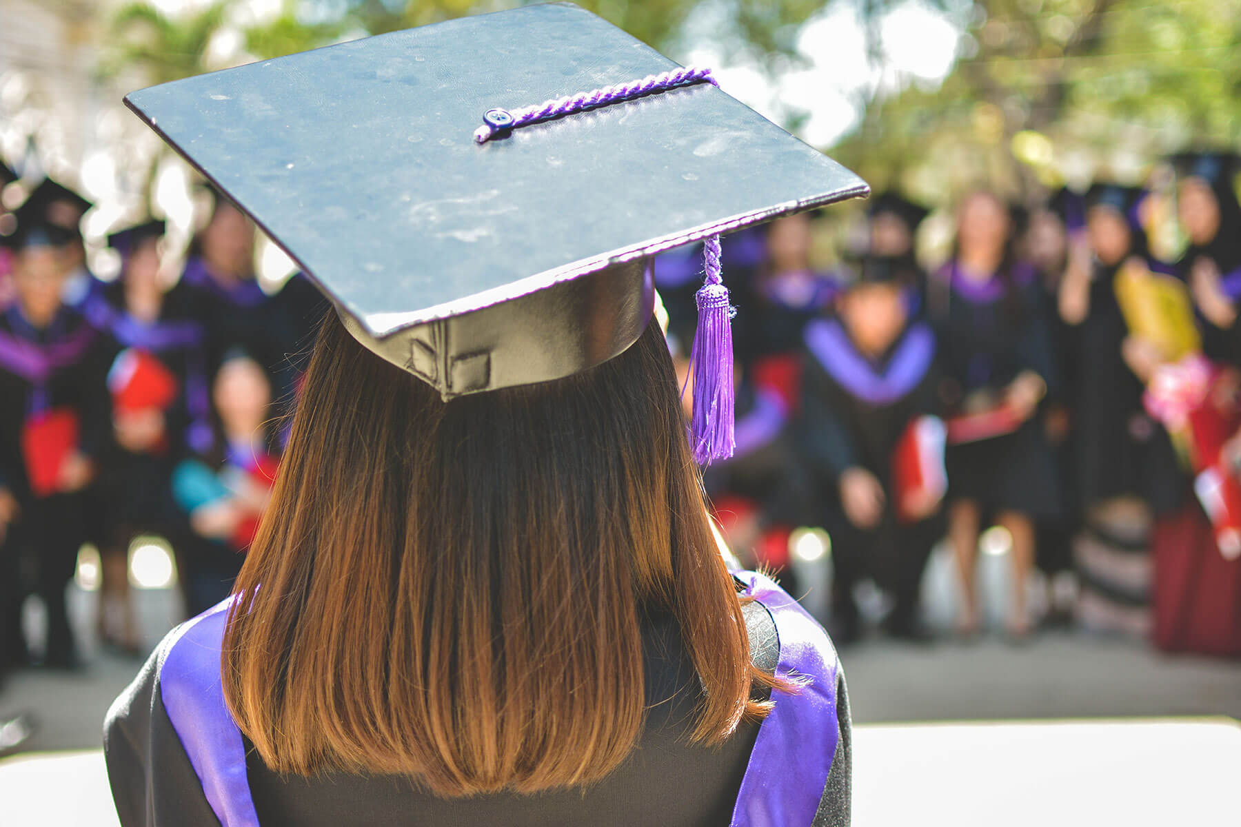 group of people in graduation gowns and caps, one girl faces away from image and looks towards group